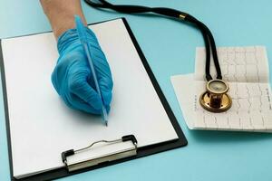 médico mão dentro uma luva escreve alguma coisa em uma tábua com uma papel com uma caneta Próximo para isto mentiras uma estetoscópio e uma cardiograma foto