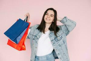 alegre comprador. jovem mulher dentro jeans camisa a comemorar, braço elevado dentro excitação, contra Rosa fundo, Deleitado de conectados compras ou surpreendente encontrar foto