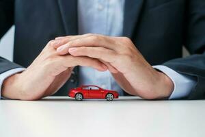 capa de mão de empresário ou brinquedo de carro vermelho de proteção na mesa. seguro de carro, garantia, reparo, financeiro, bancário e conceito de dinheiro foto