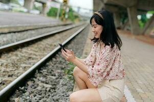 ásia jovem mulher viajante com tecelagem cesta usando uma Móvel telefone ao lado estrada de ferro trem estação dentro Bangkok. viagem viagem estilo de vida, mundo viagem explorador ou Ásia verão turismo conceito. foto