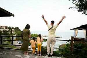 traseiro Visão do família com dois filhas em pé com braços elevado e olhando às a mar dentro nessebar. foto