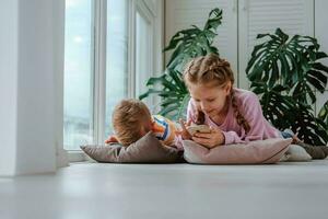 crianças mentira em a chão do a janela e Assistir desenhos animados em seus telefones. irmão e irmã jogar jogos em a telefone. foto
