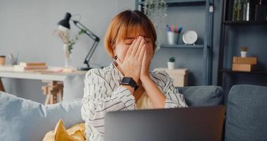 A senhora freelance asiática sente dor de cabeça enquanto está sentada no sofá com um laptop on-line, aprende na sala de estar de casa foto