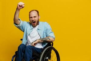 homem dentro uma cadeira de rodas gritos com raiva, mostra punho parece às a Câmera é não satisfeito, com tatuagens em dele mãos senta em amarelo fundo do a estúdio, a conceito do saúde é pessoa com deficiências foto