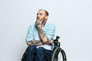 uma homem dentro uma cadeira de rodas parece às a Câmera arrogância, com tatuagens em dele braços senta em uma cinzento estúdio fundo, a conceito do saúde é uma pessoa com deficiências foto