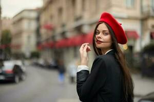 moda mulher retrato sorrir dentes em pé em a rua dentro a cidade fundo dentro à moda roupas com vermelho lábios e vermelho boina, viagem, cinematográfico cor, retro vintage estilo, urbano moda estilo de vida. foto