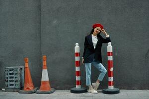 moda mulher em pé inclinado contra uma parede rua contra uma fundo do a cidade estrada trabalho turista dentro à moda roupas com vermelho lábios e vermelho boina, viagem, cinematográfico cor, retro vintage estilo. foto