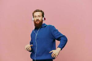 retrato do uma ruiva homem vestindo fones de ouvido sorridente e dançando, ouvindo para música em uma Rosa fundo. uma hipster com uma barba. foto