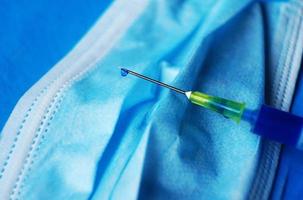 uma macro detalhada de uma seringa com uma gota de líquido azul através da agulha em uma máscara médica com fundo azul foto