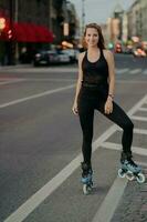 tiro de comprimento total de jovem ativa estar em boa forma física vestida de activewear preto gosta de andar de patins durante o bom dia de verão poses na estrada contra o fundo da cidade. lazer. foto