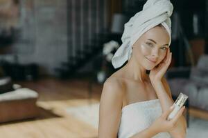 pele Cuidado conceito. mulher aplica-se face loção, embrulhado dentro toalha. diariamente mimos, facial tratamentos. nu ombros, saudável tez. foto