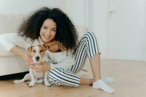 cabelo encaracolado modelo poses dentro de casa com Amado jack russell terrier. concurso abraçar, sentado em chão perto confortável sofá. estimando Tempo junto. foto