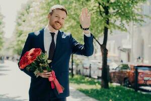 confiante homem de negocios dentro azul terno detém vermelho rosa ramalhete, esperando para esposa às parque, alegremente ondas e sorri. foto