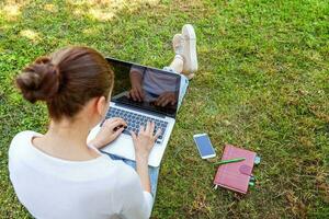 jovem mulher sentada no gramado verde no parque da cidade trabalhando no computador portátil pc. conceito de negócio freelance foto