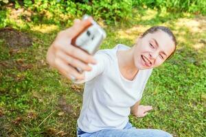 jovem menina levar selfie a partir de mãos com telefone mostrando língua e engraçado face sentado em parque ou jardim fundo foto