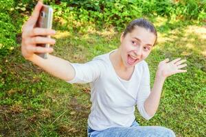 jovem engraçado menina levar selfie a partir de mãos com telefone sentado em verde Relva parque ou jardim fundo foto