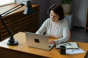 confiante à moda europeu meio envelhecido Senior mulher usando computador portátil às lar. à moda Mais velho maduro anos 60 senhora sentado às mesa olhando às computador tela digitando conversando lendo escrevendo e-mail. foto