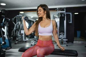 lindo Atlético muscular mulher bombas acima a músculos de 1 braço elevadores haltere exercício em Banco dentro ginástica academia. jovem esporte menina ganhos Forte fisica músculo bem de peso levantado dentro ginástica estúdio foto