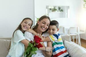 jovem mãe com uma ramalhete do rosas risos, abraçando dela filho, e alegre menina com uma cartão parabeniza mãe durante feriado celebração dentro cozinha às casa foto