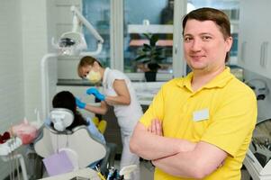 confiante feliz masculino dentista dentro amarelo uniforme em pé dentro dental escritório com braços guardada, sorridente olhando às Câmera foto