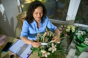 fechar-se inspirado mulher, florista, evento Gerente colocando uma cumprimento cartão dentro uma floral arranjo dentro a vime cesta foto
