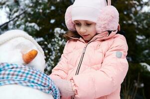 fechar-se adorável criança menina dentro Rosa baixa Jaqueta e fofo protetores de ouvido, colocando em uma azul cachecol por aí a boneco de neve pescoço foto