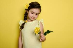 tímido bebê menina com amarelo rosas dentro dela Penteado, olhando às uma lindo rosa flor, isolado em estúdio fundo foto