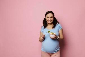 isolado retrato em Rosa fundo do uma lindo grávida mulher com uma tigela do fruta, pronto para comer. saudável nutrição foto