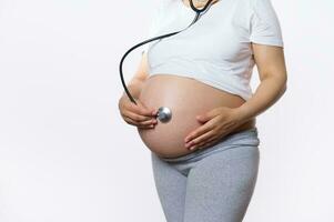 fechar-se grávida mulher coloca uma estetoscópio em dela barriga e escuta para batimento cardiaco do dela bebê dentro útero. feto desenvolvimento foto