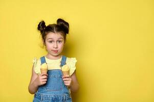 feliz pequeno criança menina mostrando dela língua às Câmera, segurando gostoso gelo creme dentro waffle cones, isolado amarelo fundo foto