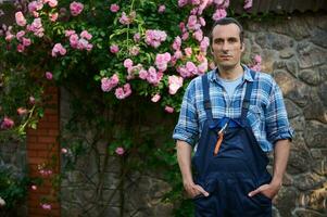 bonito masculino jardineiro dentro jardinagem roupa, olhando às Câmera, em pé às dele local de trabalho dentro a quintal do uma mansão foto