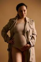 atraente confiante grávida mulher dentro lingerie e trincheira casaco, acariciando dela barriga, isolado em bege estúdio fundo foto