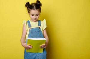 retrato do fofa criança menina dentro amarelo camiseta e azul jeans vestir macacão, segurando livro didático isolado estúdio fundo foto