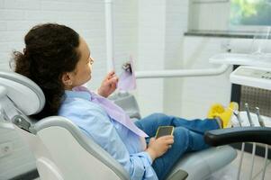 fêmea paciente sentado dentro Dentistas cadeira, olhando dentro a espelho depois de profissional dental tratamento dentro odontologia clínica foto