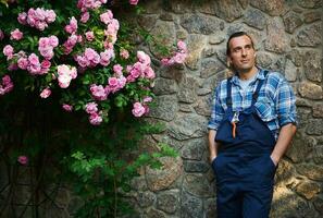 caucasiano bonito jardineiro anos 40, dentro trabalhos uniforme, em pé com mãos dentro bolsos perto florescendo rosas arbusto, olhando a parte, de lado foto