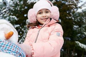 autêntico lindo feliz pequeno criança menina jogando com uma boneco de neve em uma Nevado inverno parque, sorridente olhando às Câmera foto