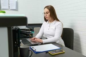 recepcionista trabalho em computador portátil, sentado atrás uma recepção escrivaninha dentro moderno clínica, guardando compromissos com médico funcionários foto