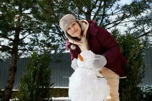 lindo alegre jovem mulher dentro protetores de ouvido chapéu faz uma boneco de neve, tendo Diversão dentro uma neve coberto natureza. inverno lazeres foto