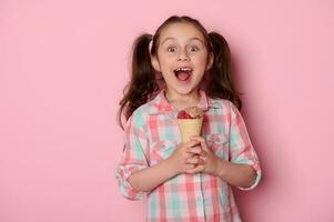 estúdio retrato em Rosa fundo do uma lindo espantado pequeno criança menina com uma saboroso gelo creme dentro waffle cone foto