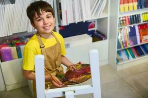 sorridente adolescente Garoto artista, segurando paleta com óleo tintas, pintura sentado às a cavalete contra a telas fundo foto