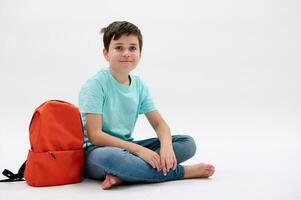 inteligente adolescente Garoto dentro azul camiseta e jeans com laranja mochila, sorrisos olhando às Câmera, isolado branco estúdio pano de fundo foto