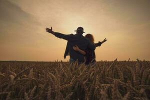 feliz homem e mulher estão em pé dentro seus agrícola campo dentro pôr do sol. elas estão cultivar trigo e desfrutando Boa agrícola temporada. foto