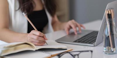 close-up de mulher asiática escrevendo no caderno em uma mesa com laptop, garota trabalha em uma cafeteria, conceito de negócio freelance foto