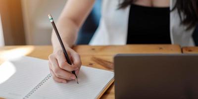 close-up das mãos de uma mulher com computador laptop, caderno e caneta fazendo anotações no escritório de negócios foto
