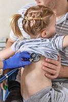 menina nos braços de seu pai no consultório médico na clínica. o médico examina a criança, ouve os pulmões com um estetoscópio. tratamento e prevenção de infecções respiratórias.