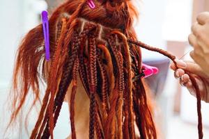 mãos de cabeleireiro trançam dreadlocks ruivos de menina. foto