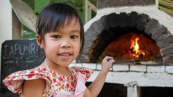 uma garotinha faz pizza feita à mão e fica sorrindo em frente a um forno de pizza de tijolos de madeira em um pátio ao ar livre em um restaurante. foto
