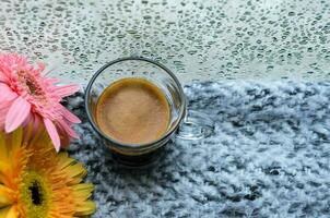 uma vidro do espresso café tiro coloca em cama com gerbera flores quando chuva solta em janela dentro chovendo dia. foto