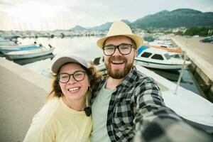 jovem casal levando uma auto retrato rindo Como elas pose fechar juntos para Câmera em seus Smartphone ao ar livre dentro verão porta marina com barcos e iates foto