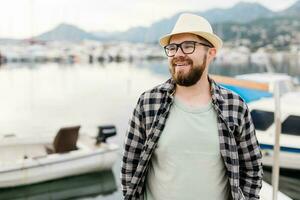 bonito homem vestindo chapéu e óculos perto marina com iates. retrato do rindo homem com mar porta fundo com cópia de espaço foto